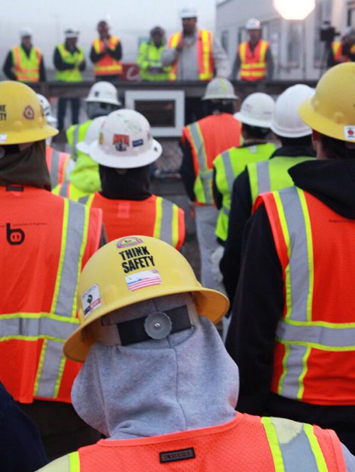 A group of workers in high vis and hard hats are listening to a presentation