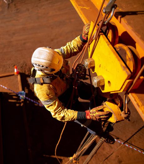 View from above of a man working on mining equipment in a harness. The background is red dirt.