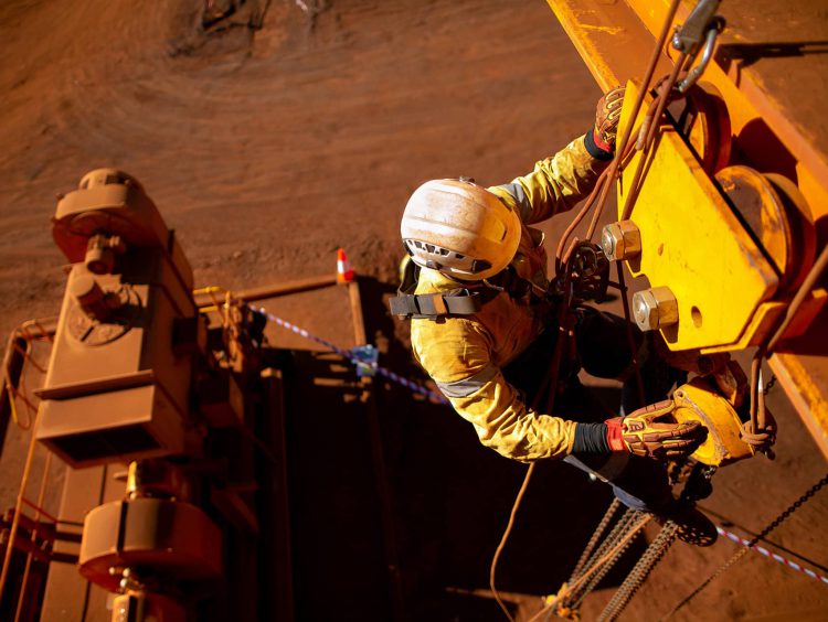 View from above of a man working on mining equipment in a harness. The background is red dirt.